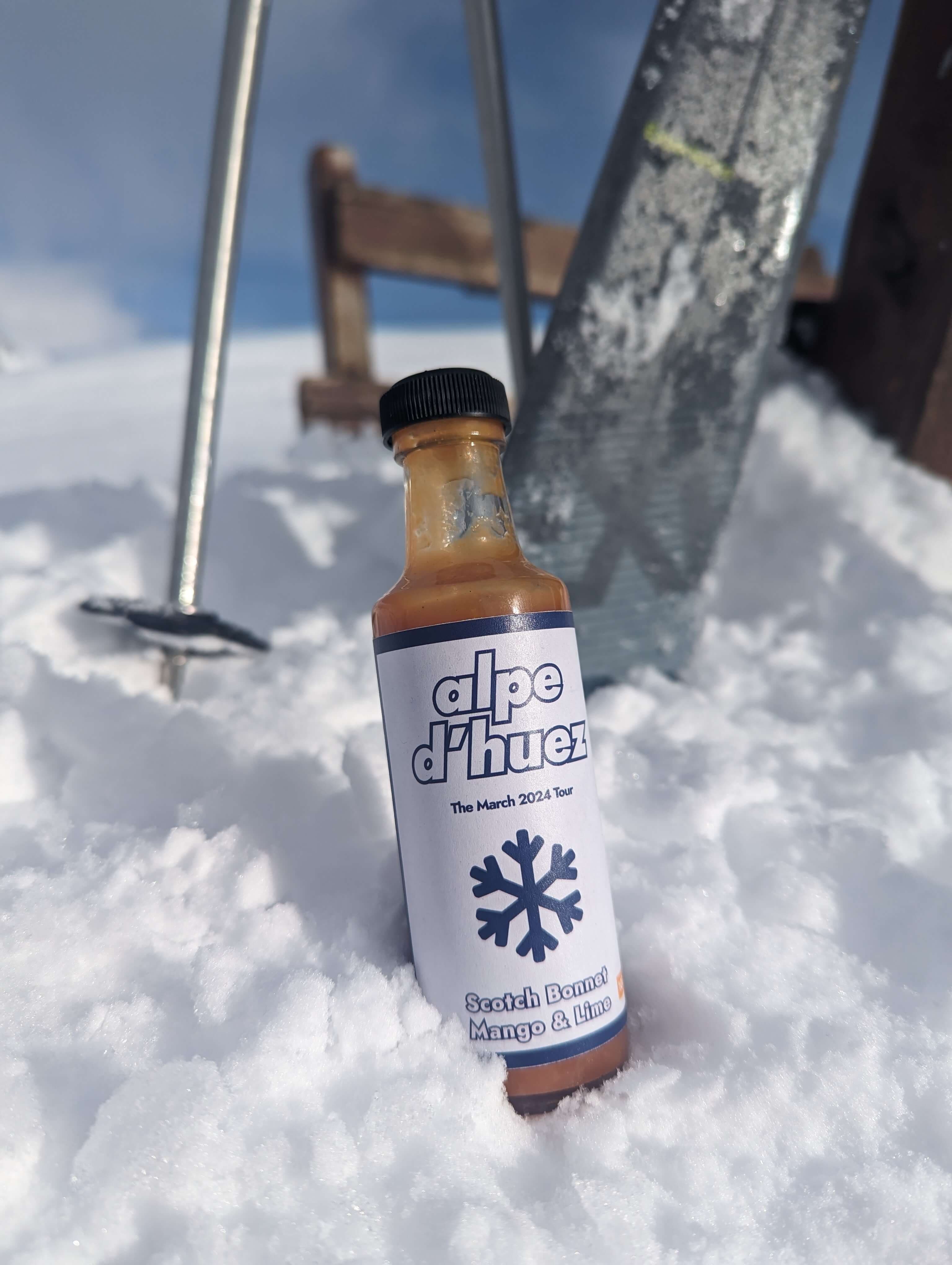 A closeup of a custom branded bottle of hot sauce. The bottle is on the ground in the snow. Behind it are skis and ski poles